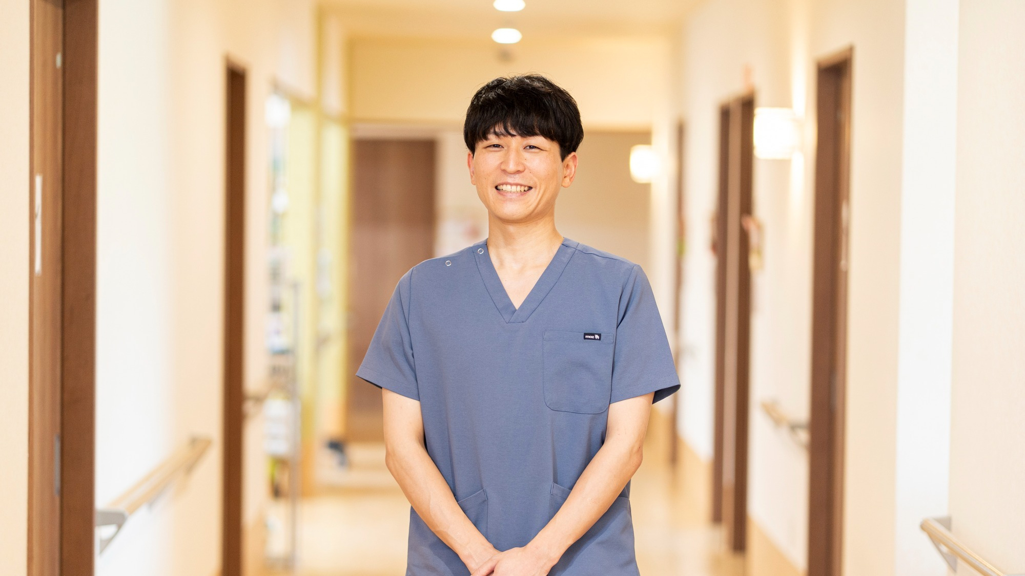 廊下に立つ介護職員の男性。青色の医療用ユニフォーム着用。